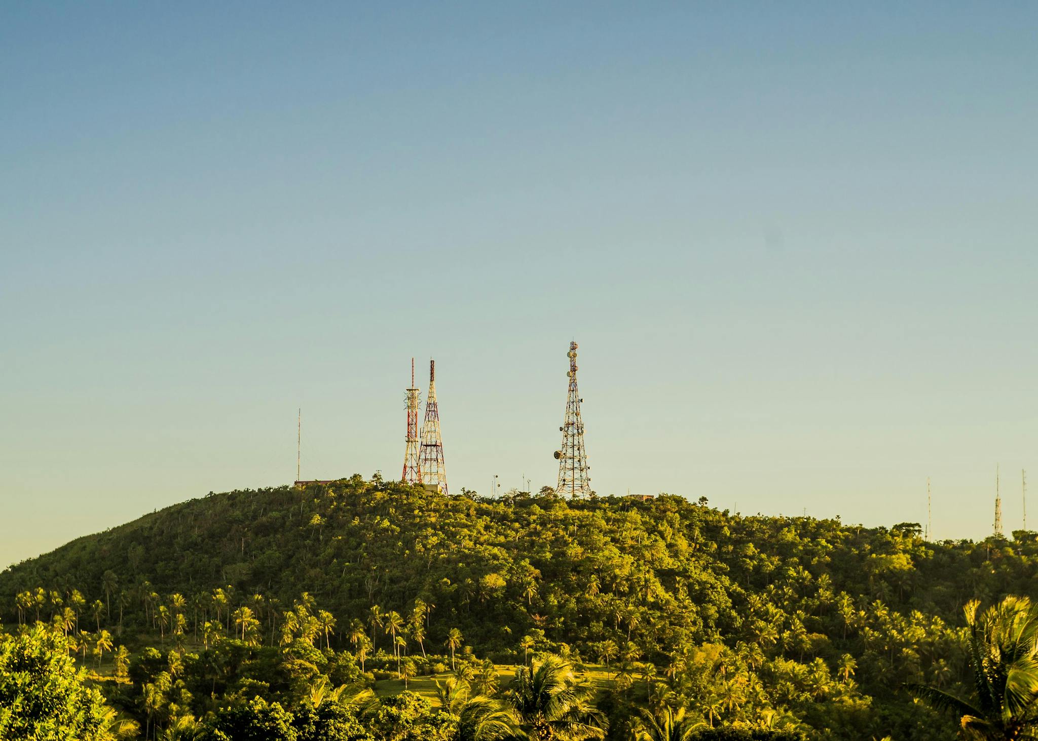 Metal tall telecommunication towers on high hills covered with tropical trees with fresh verdant foliage under cloudless sky